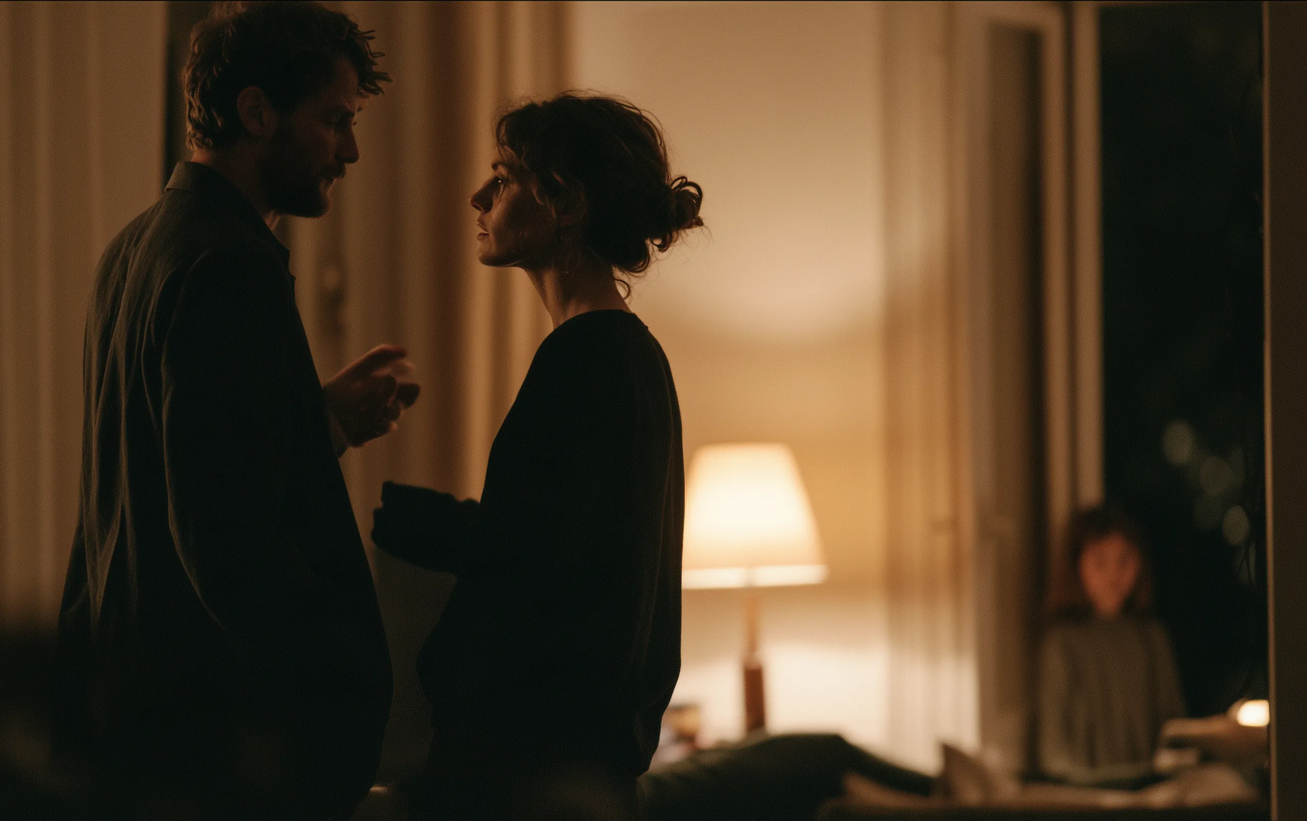 Two adults standing close and facing each other in a lamplit living room, one of them with a hand half raised; at the edge of the frame a child stands in the doorway watching them