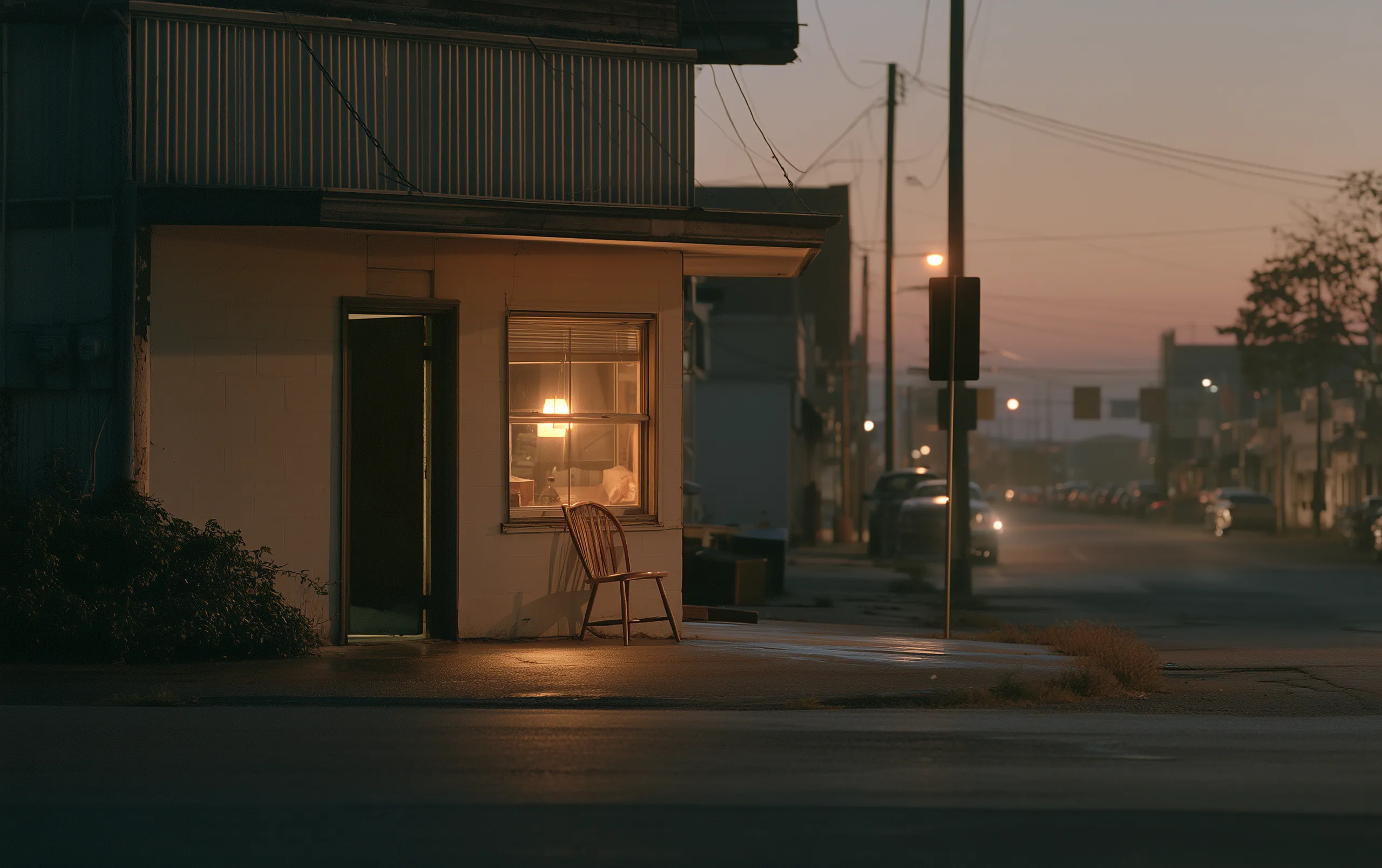 An ordinary street corner in the last light, a lit window, a chair somebody left outside, nobody there